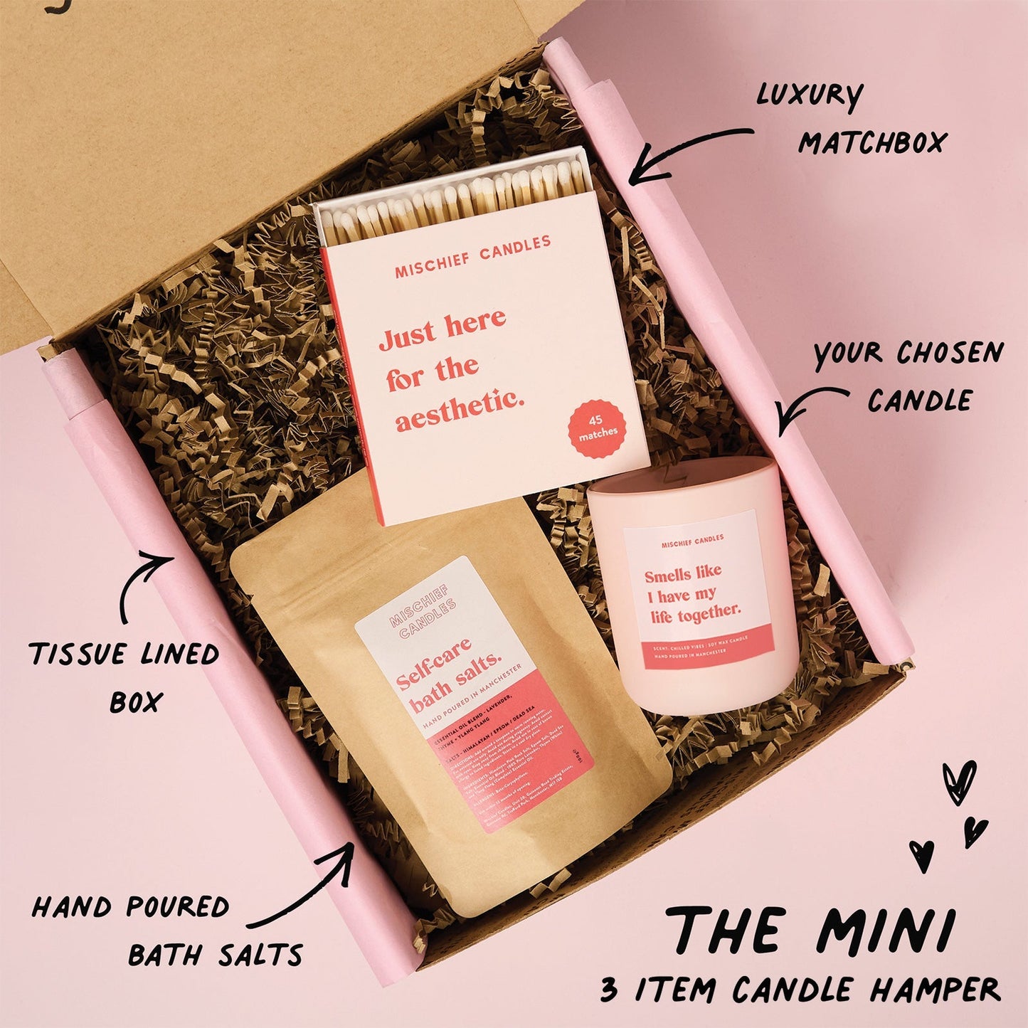 Candle hamper with matchbox, bath salts, and candle in a tissue-lined box on a pink background.