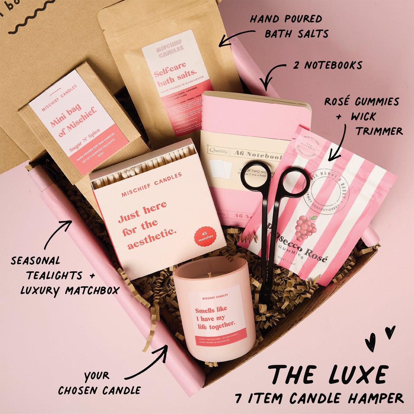 Candle hamper with various items including candles, notebooks, and matchbox on a pink background.