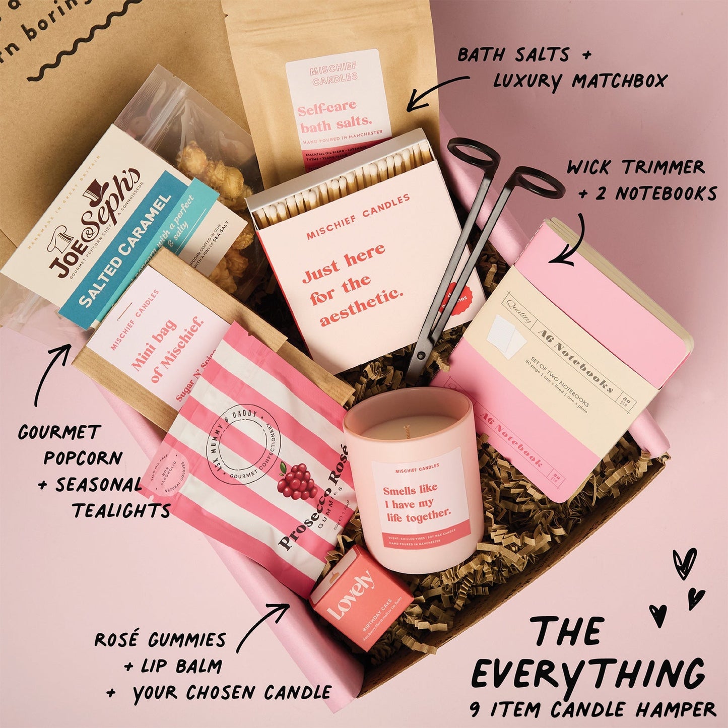 Candle hamper with various items including a candle, lip balm, and bath salts on a pink background.