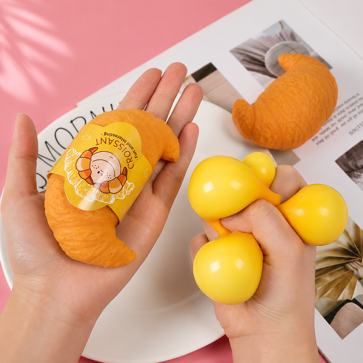 Hands holding a small orange plush toy and a yellow dumbbell-shaped stress reliever on a magazine background.