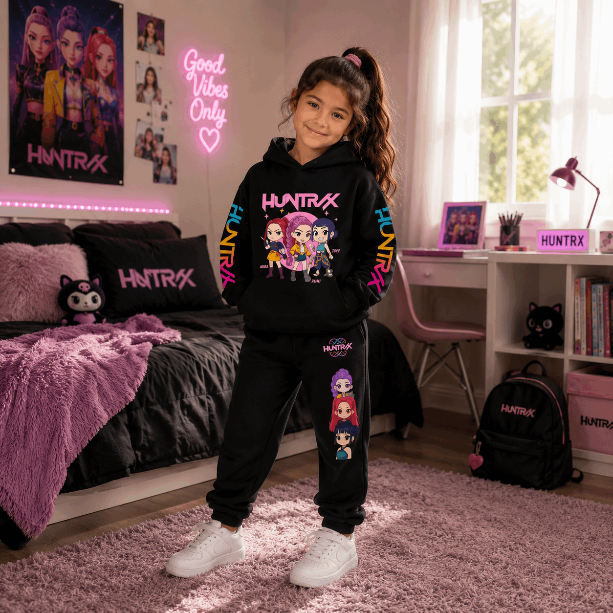 A young girl stands in a pink bedroom wearing a black HUNTRX hoodie and matching black sweatpants with colorful cartoon character graphics and pink lettering. The room includes a bed, desk, window, and HUNTRX-branded items.