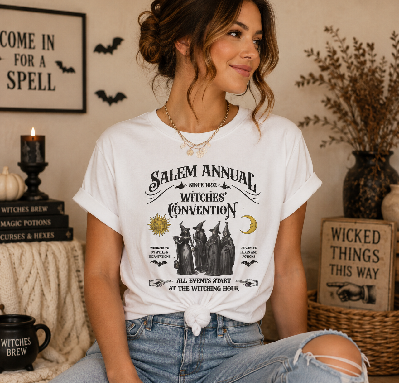 A woman wearing a white graphic t-shirt with a 'Salem Annual Witches Convention' print, styled in a cozy, decorated room with Halloween-themed decor.