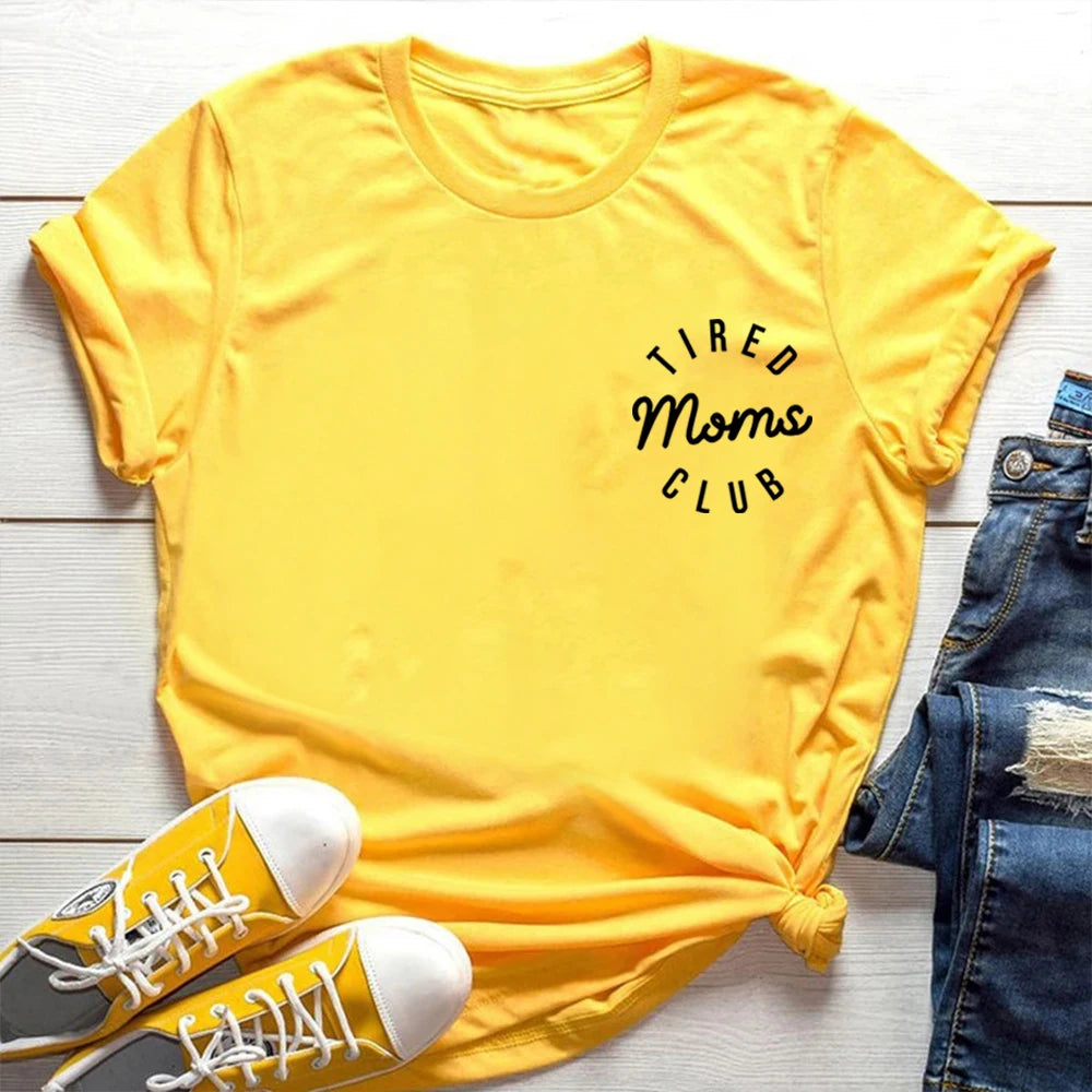Yellow t-shirt with 'Tired Moms Club' text, paired with yellow sneakers and blue jeans on a light wooden surface.