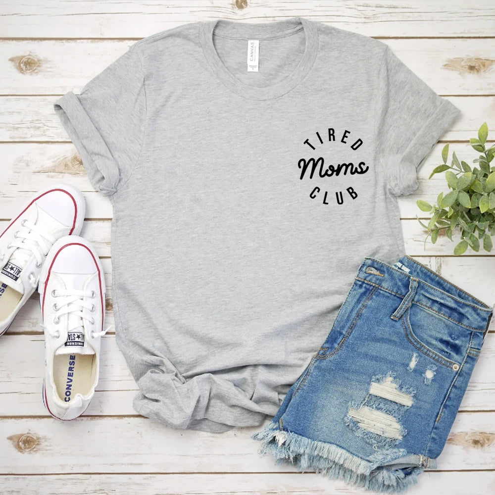 Gray t-shirt with 'Tired Moms Club' text, white sneakers, and denim shorts on a wooden surface.