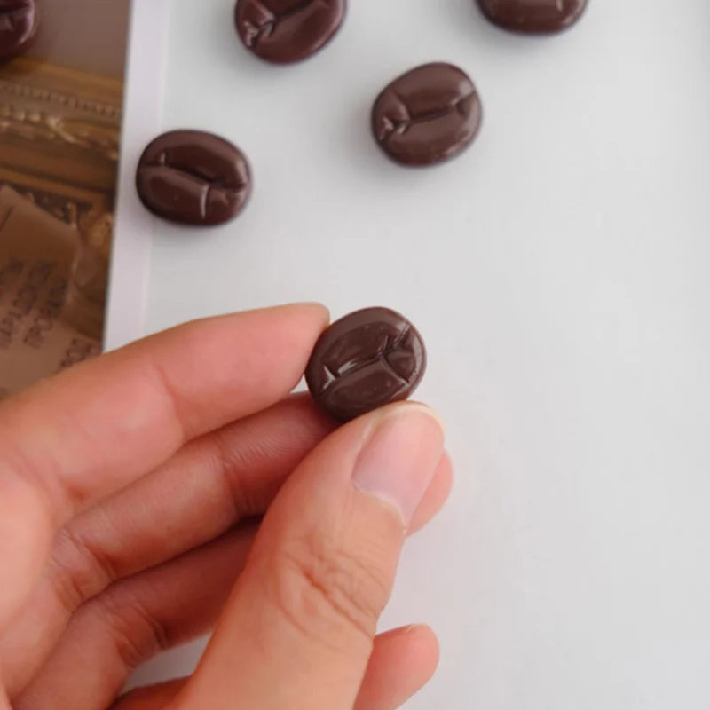coffee bean magnet held between fingers with more beans on a white surface
