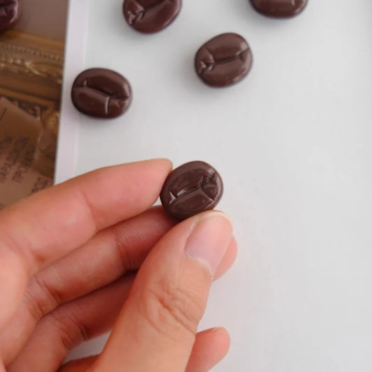 coffee bean magnet held between fingers with more beans on a white surface