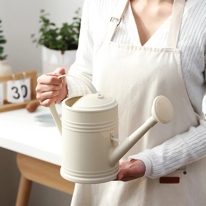 Person holding a white watering can in a home setting with plants.