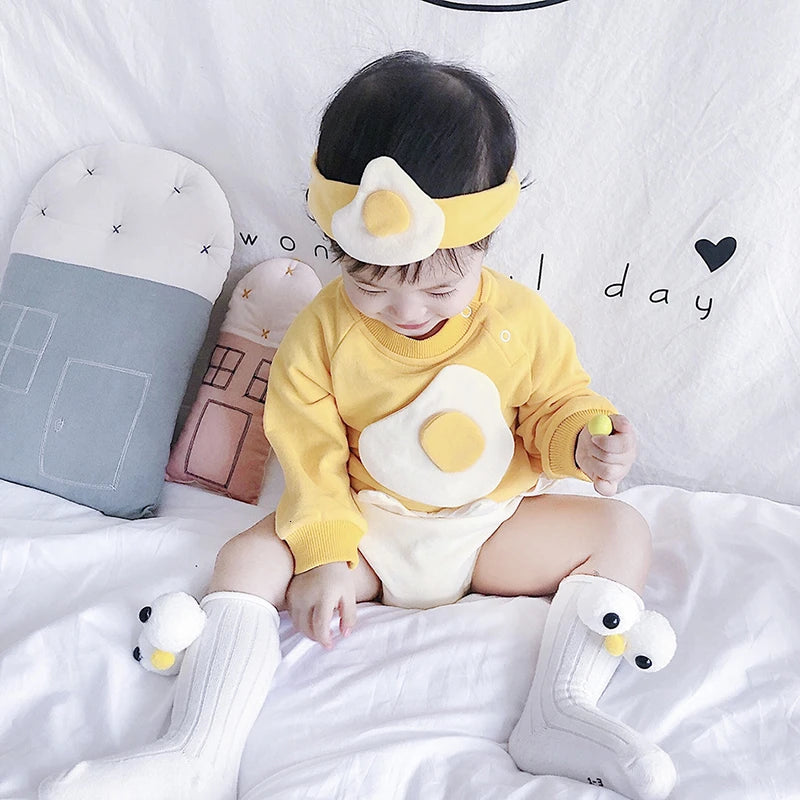 Baby in a yellow and white outfit with swan boots and headband, sitting on a bed.