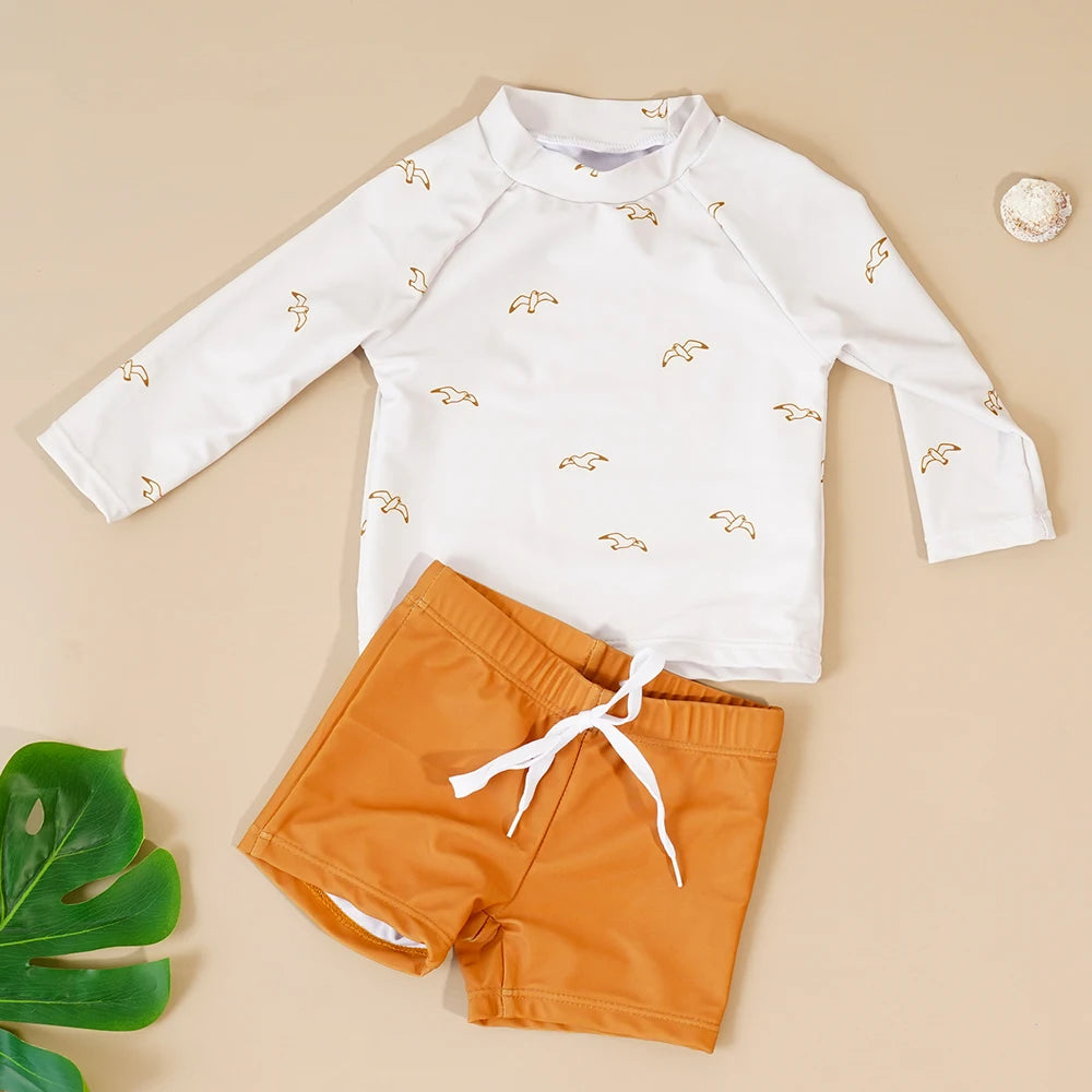 Children's white long-sleeve shirt with gold bird patterns and orange shorts on a beige background.