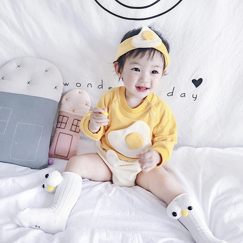 Baby in a yellow outfit with a cartoon design, sitting on a white surface with decorative pillows and toys.