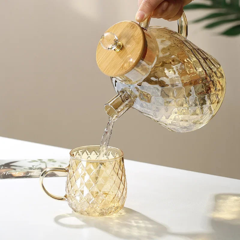 Glass teapot with wooden lid being poured into a matching glass mug on a neutral background