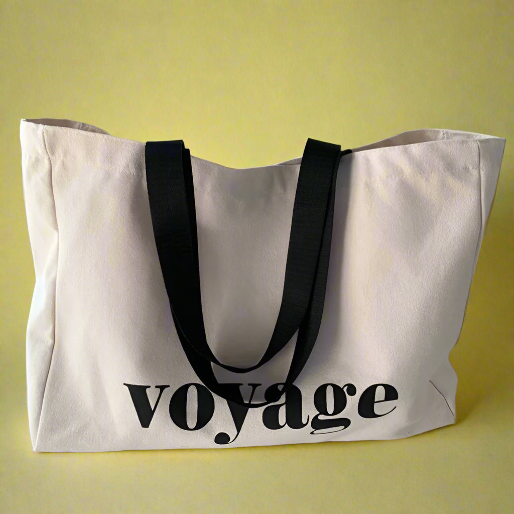 Beige tote bag with 'voyage' text and black handles on a light surface.