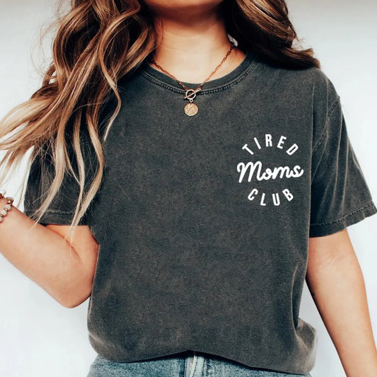 Person wearing a dark gray t-shirt with 'Tired Moms Club' text on a plain background