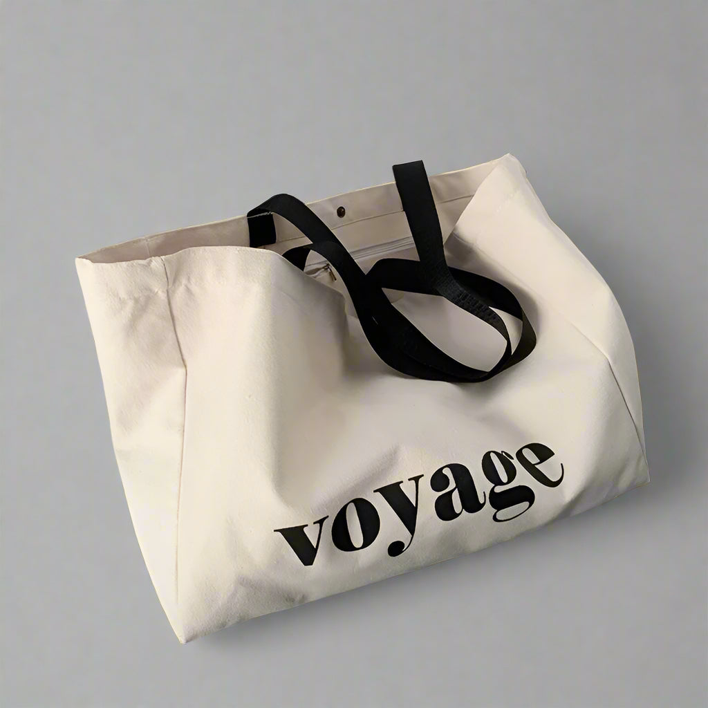 Beige tote bag with 'voyage' text on a gray floor against a white wall