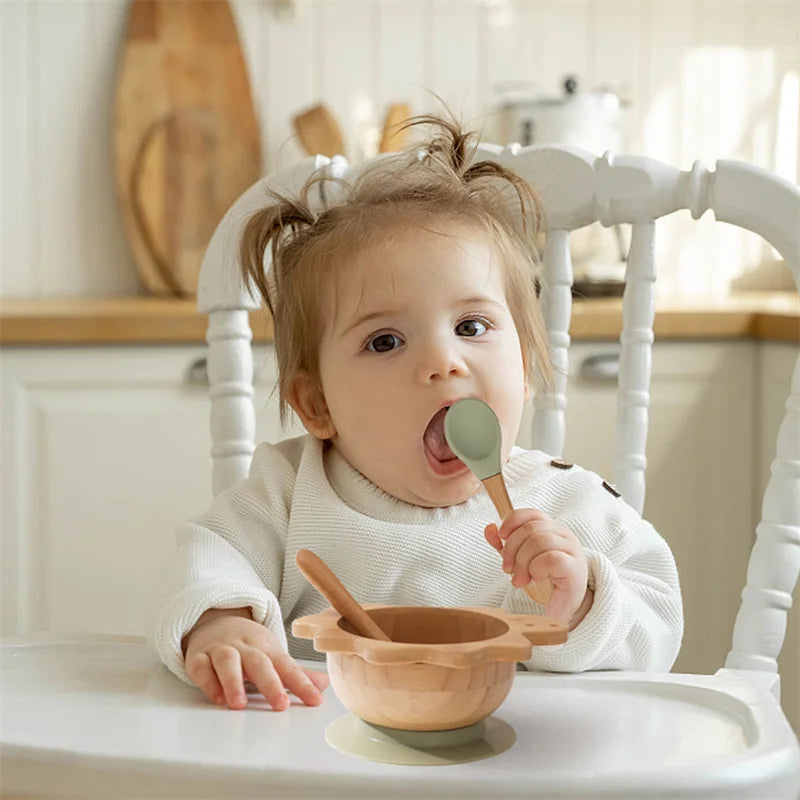 Child in a high chair eating from a wooden bowl with a green spoon in a kitchen setting.