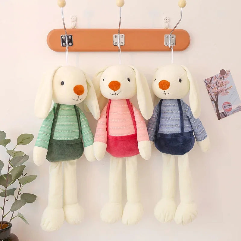 Three plush toy rabbits in colorful outfits hanging on a wooden rack against a light background.