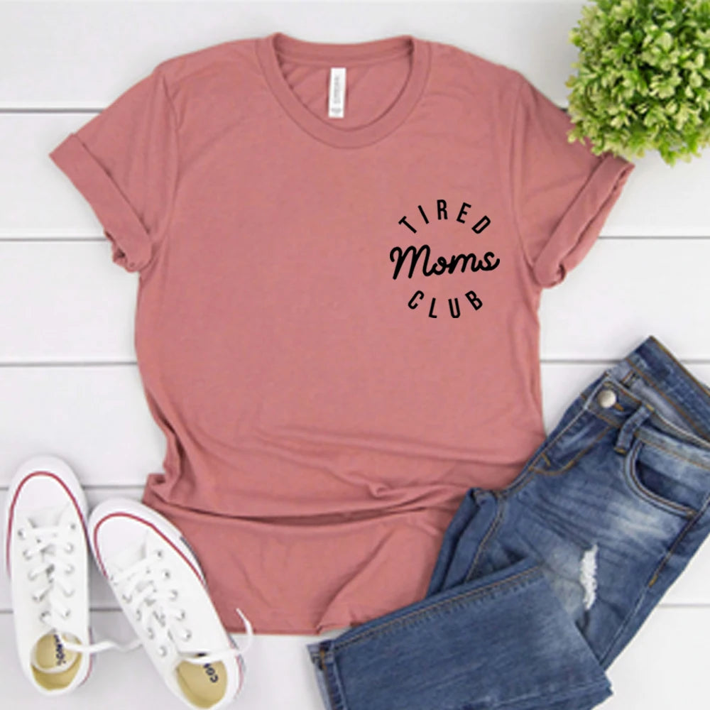 Mauve t-shirt with 'Tired Moms Club' text, paired with jeans and sneakers on a white wooden surface.