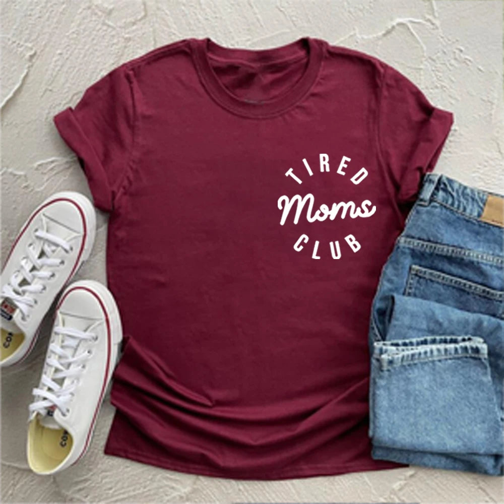 Maroon t-shirt with 'Tired Moms Club' text, paired with white sneakers and blue jeans on a light gray background.