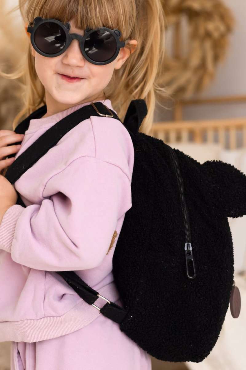 Child wearing a pink outfit and black sunglasses, carrying a black fluffy backpack.