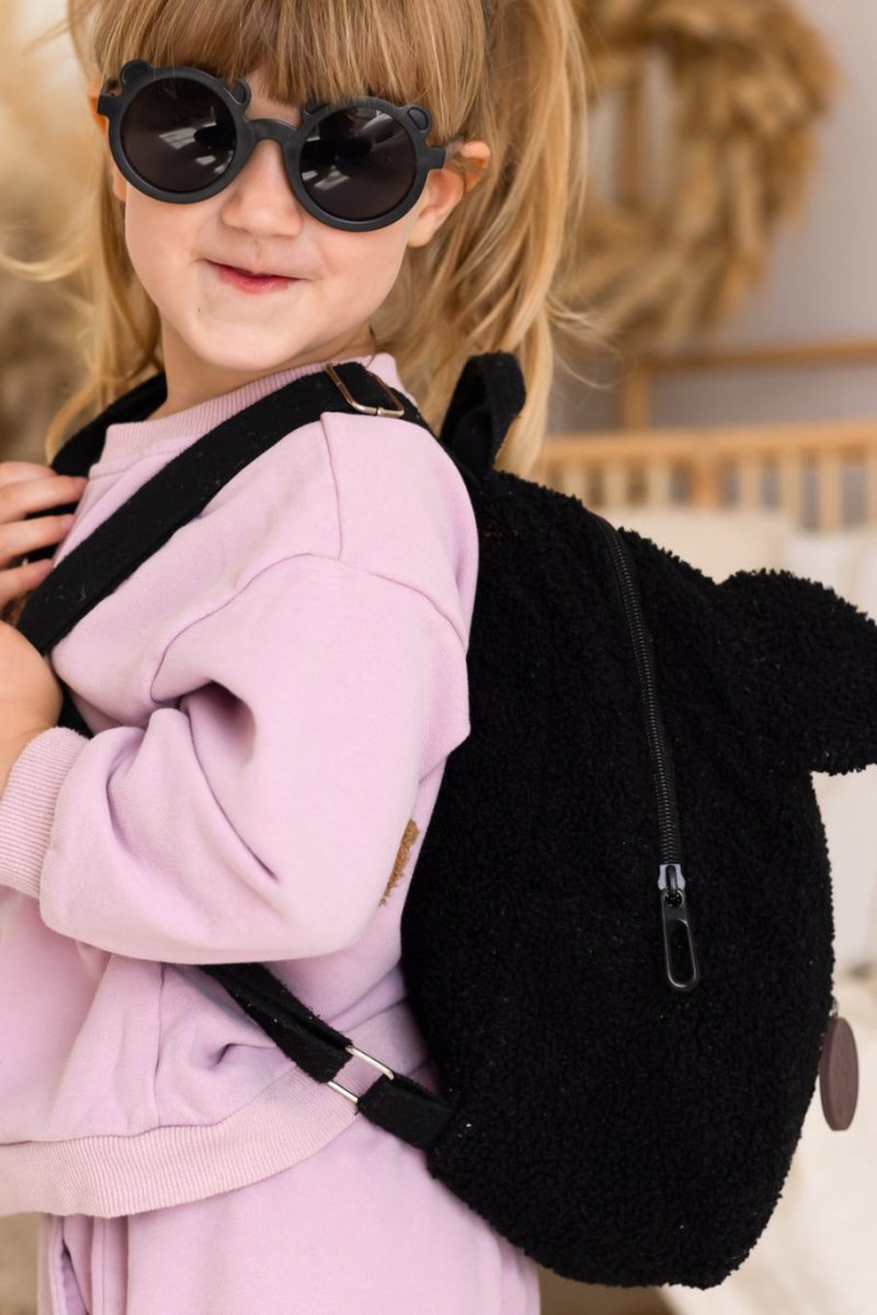 Child wearing a pink outfit and black sunglasses, carrying a black fluffy backpack.