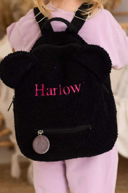 Black backpack with fluffy texture and 'Harlow' embroidered on it, worn by a person in a pink outfit.