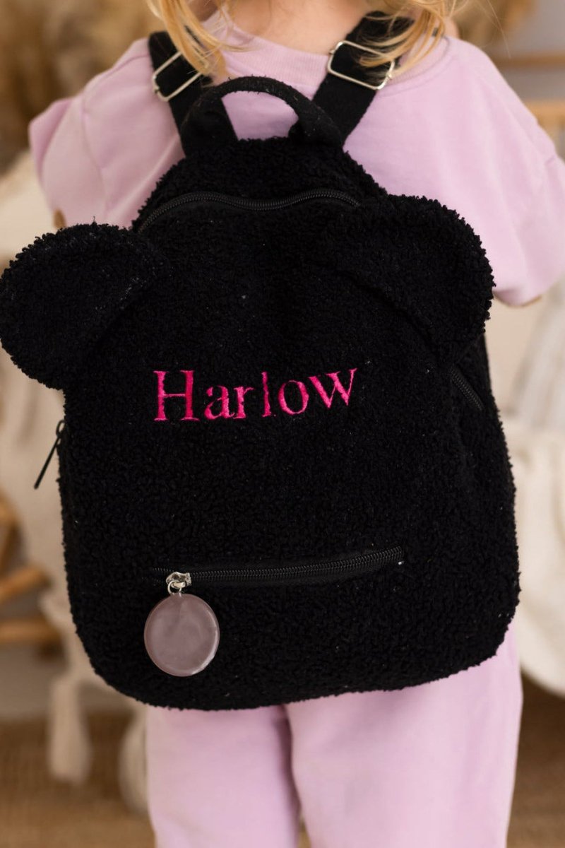Black backpack with fluffy texture and 'Harlow' embroidered on it, worn by a person in a pink outfit.