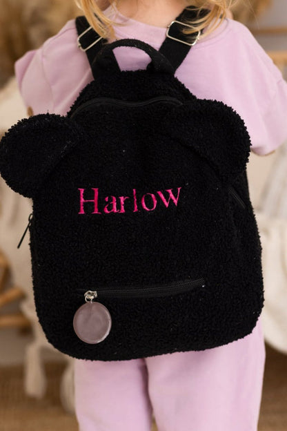 Black backpack with fluffy texture and 'Harlow' embroidered on it, worn by a person in a pink outfit.