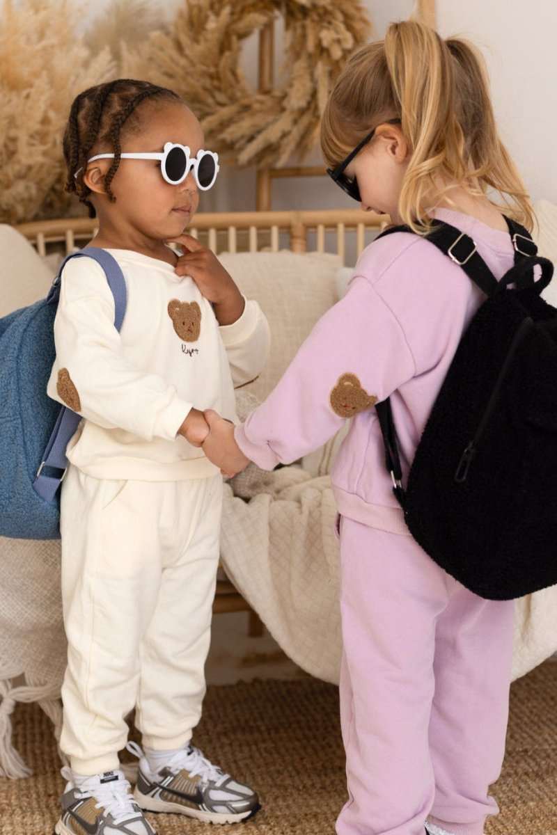 Two children in matching outfits with backpacks, standing close to each other indoors.