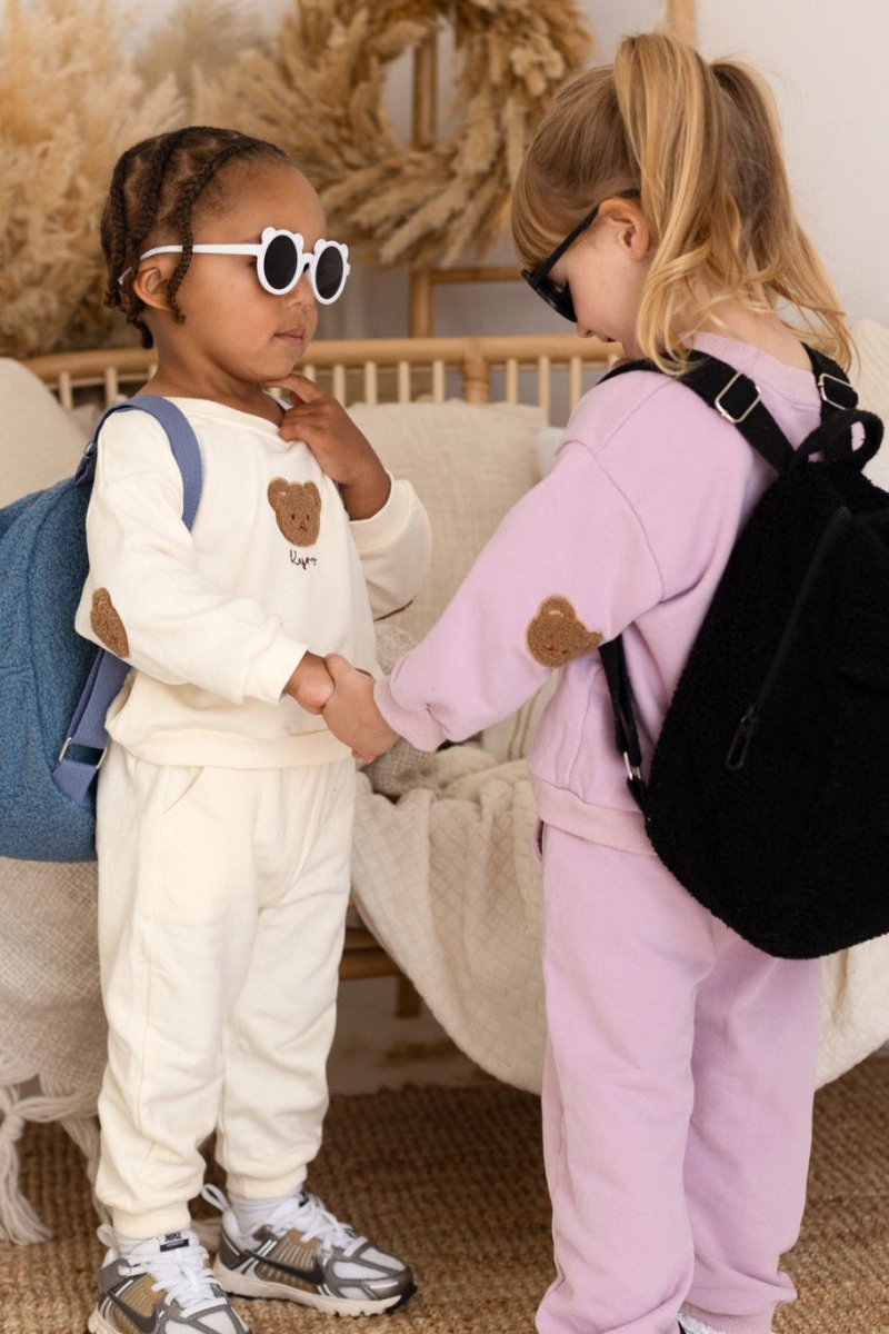 Two children in matching outfits with backpacks, standing close to each other indoors.