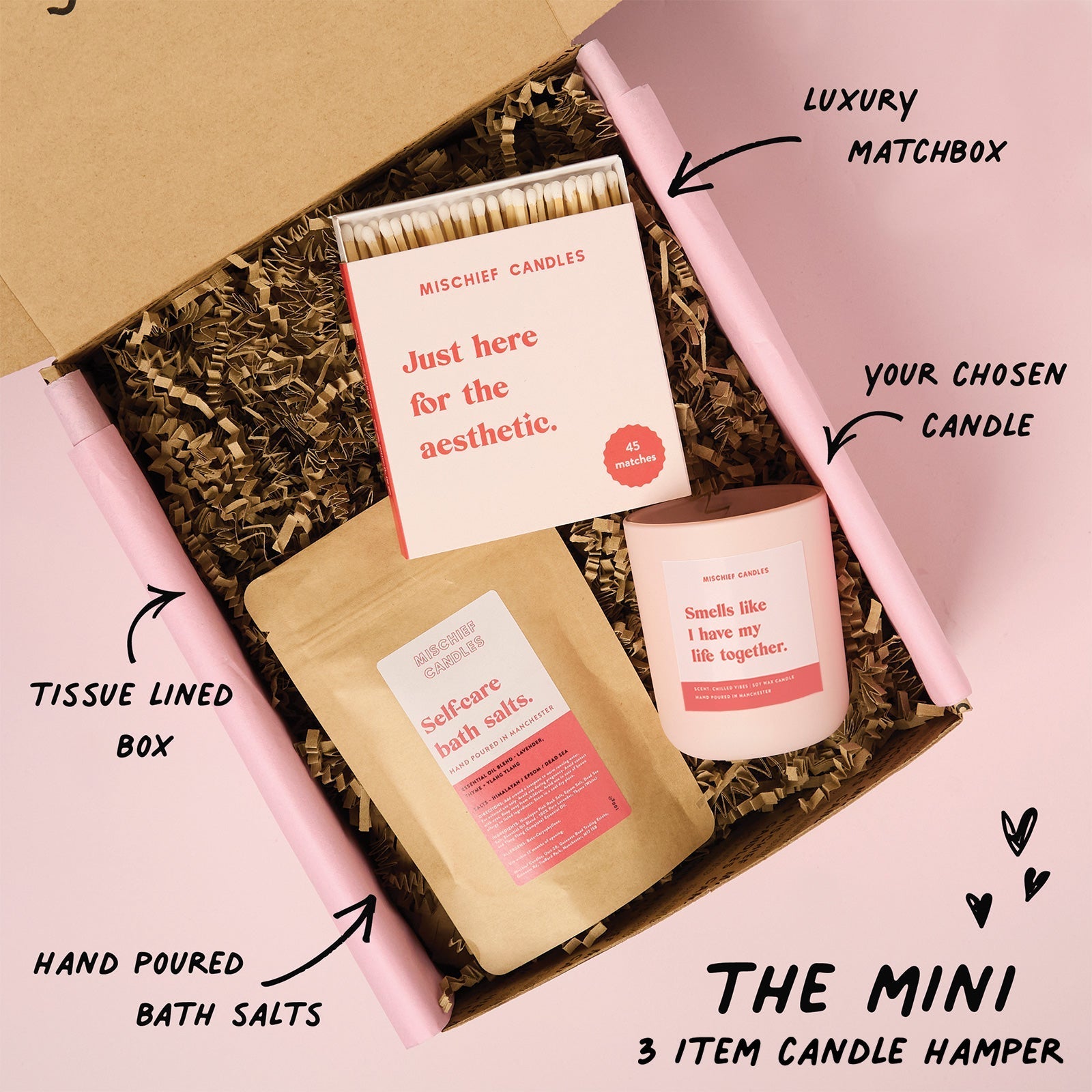 Candle hamper with matchbox, bath salts, and candle in a tissue-lined box on a pink background.
