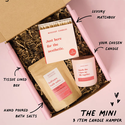 Candle hamper with matchbox, bath salts, and candle in a tissue-lined box on a pink background.