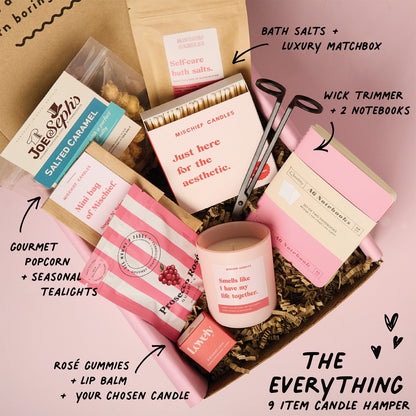 Candle hamper with various items including a candle, lip balm, and bath salts on a pink background.