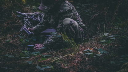 Person in camouflage gear crouching in a forest setting with a bow and arrow.