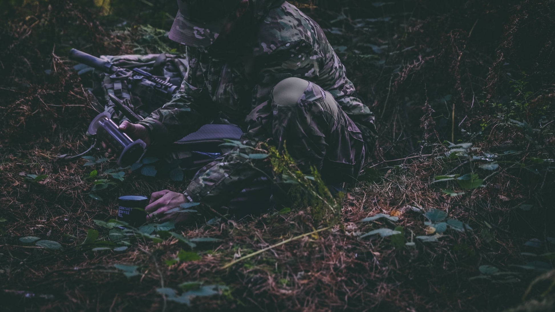 Person in camouflage gear crouching in a forest setting with a bow and arrow.