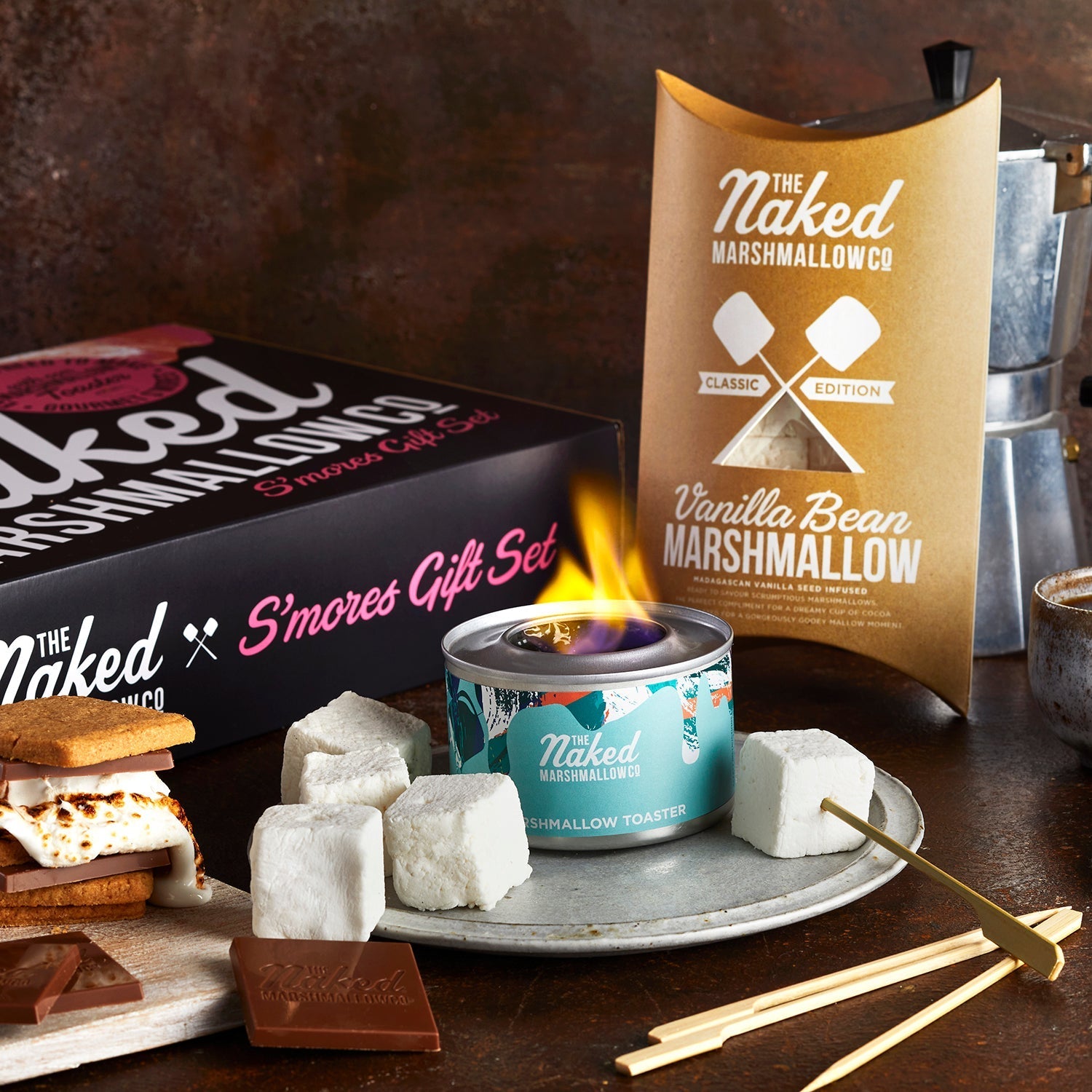 Naked Marshmallow Co. gift set with marshmallows, toasting equipment, and s'mores ingredients on a wooden surface.