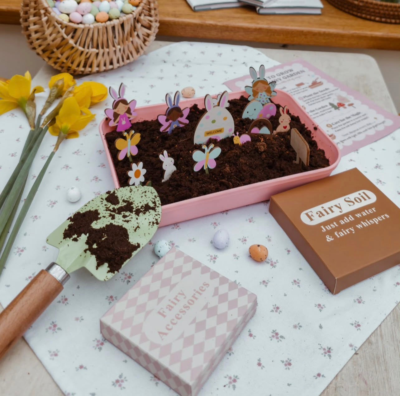 Fairy garden kit with soil, figurines, and tools on a floral tablecloth.