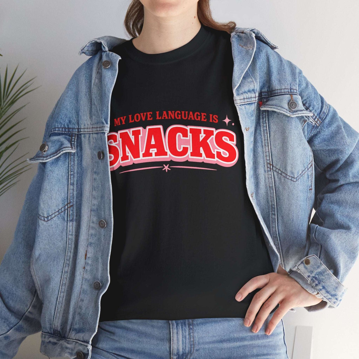 A person wearing a black graphic T-shirt with red text reading 'MY LOVE LANGUAGE IS SNACKS' under an open light-wash denim jacket, paired with blue jeans.