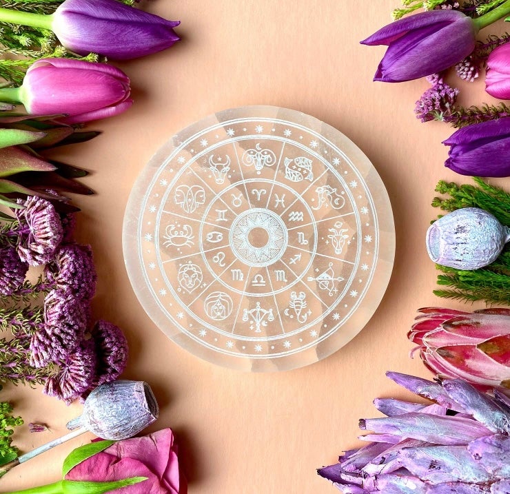 Decorative circular object with astrological symbols surrounded by purple and pink flowers on a beige background