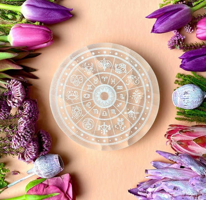 Decorative circular object with astrological symbols surrounded by purple and pink flowers on a beige background