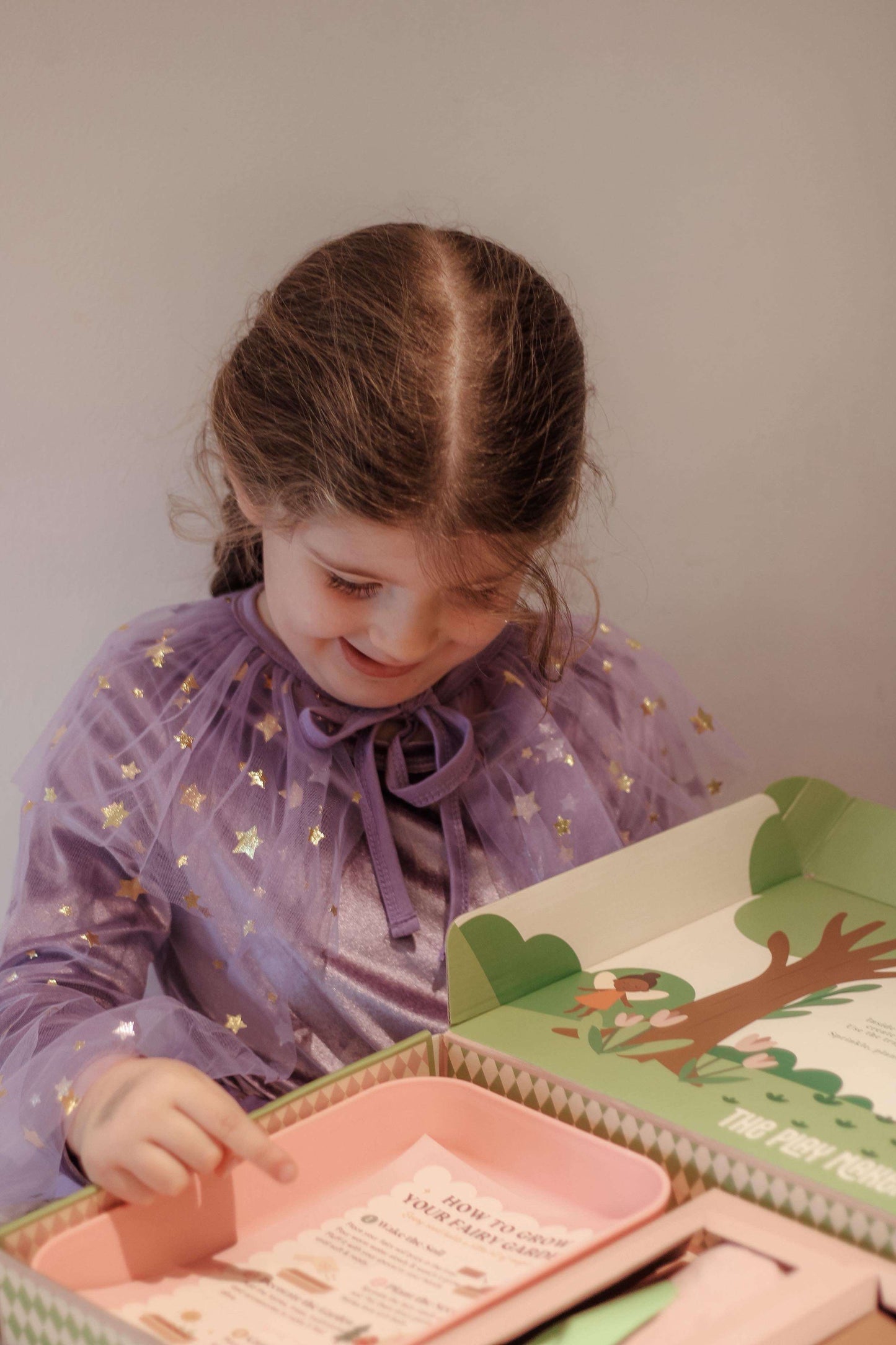 Young girl in a purple dress looking at a book with a tree illustration on a plain background