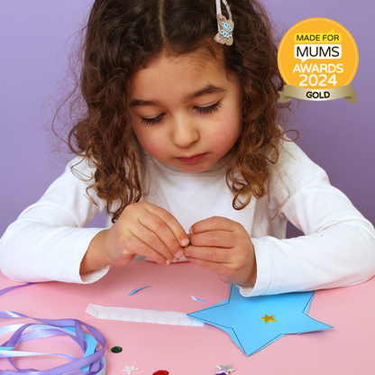 Child engaged in craft activity with decorative items on a pink table, against a purple background, with 'Made for Mums Awards 2024 Gold' badge.