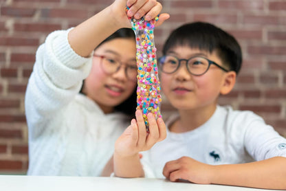 Two children playing with colorful slime in front of a brick wall.