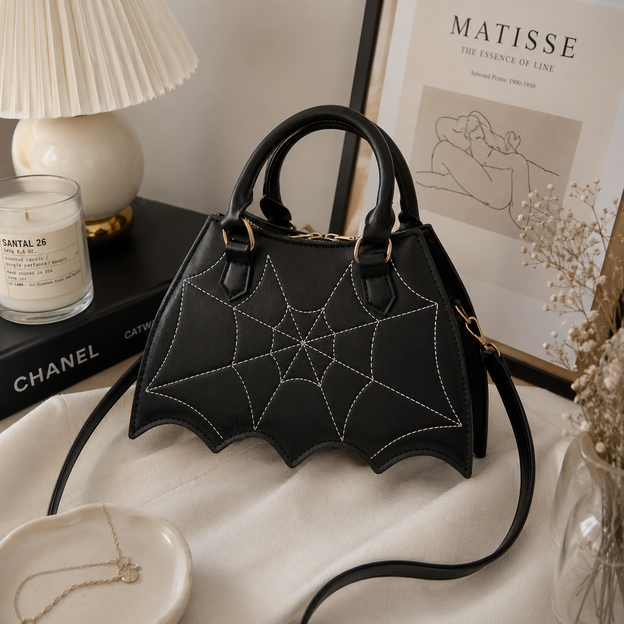 Black faux-leather handbag with a bat-wing silhouette and white spiderweb embroidery, styled on a cream fabric surface with a lamp, candle, framed print, and jewelry nearby.