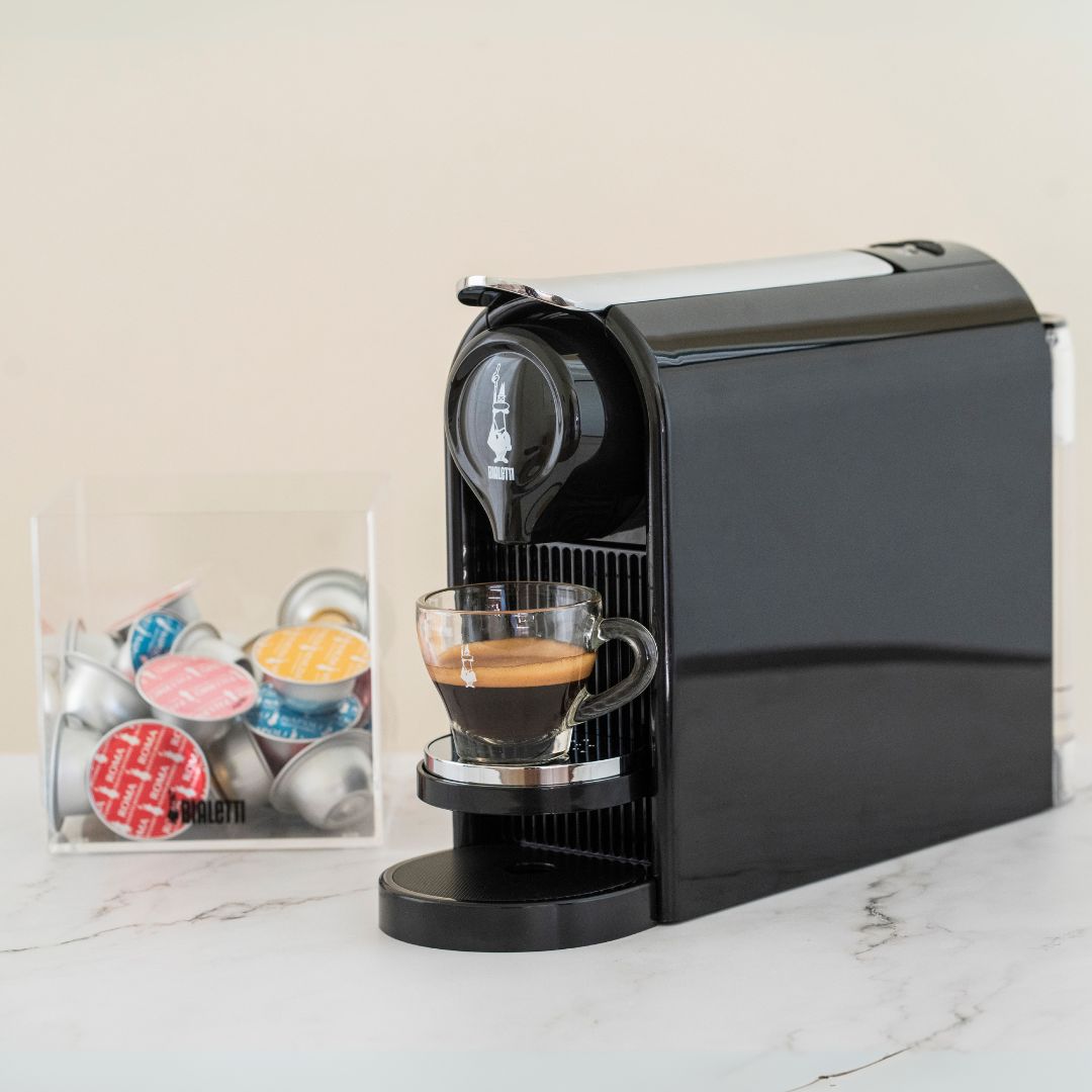Black coffee machine dispensing a cup of coffee on a marble surface with colorful capsules in the background.