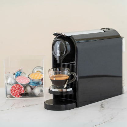 Black coffee machine dispensing a cup of coffee on a marble surface with colorful capsules in the background.