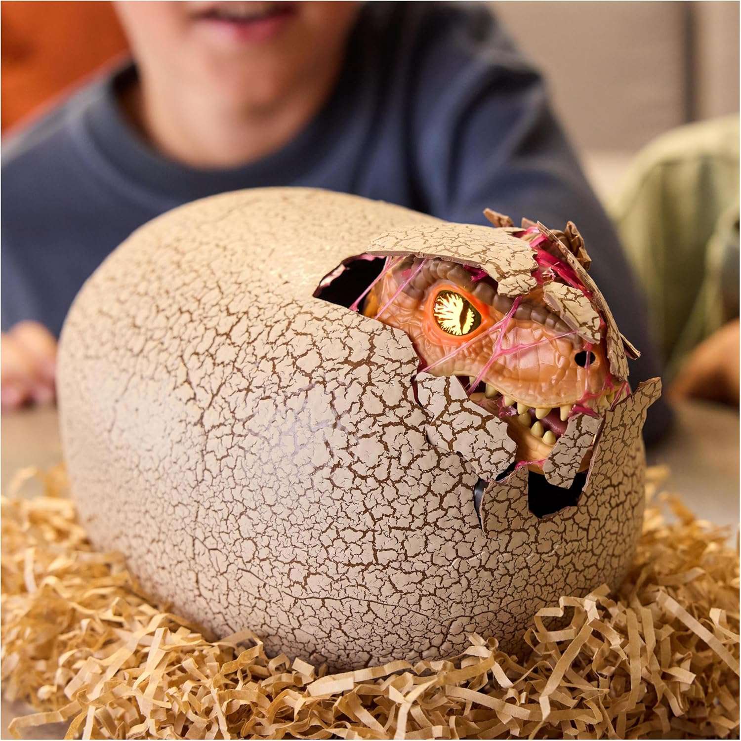 Toy dinosaur hatching from a cracked egg on a bed of straw with a child in the background.
