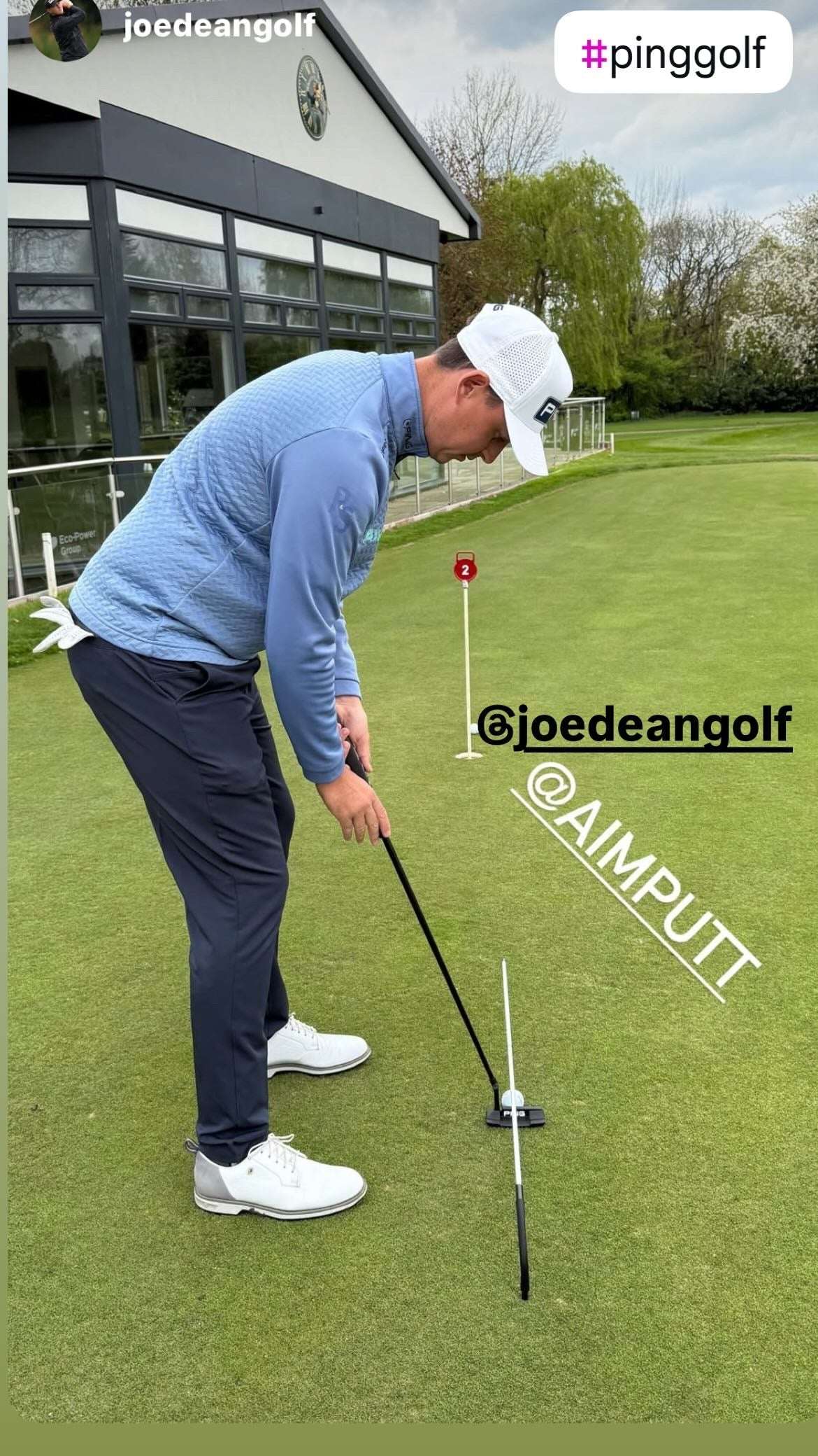 Man practicing golf on a green course with a building and trees in the background.