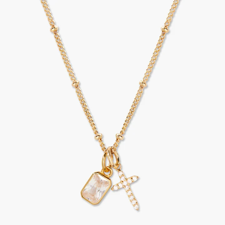 A gold chain necklace featuring a rectangular bezel-set pale pink stone and a small gold cross charm paved with clear stones, shown on a light gray background.