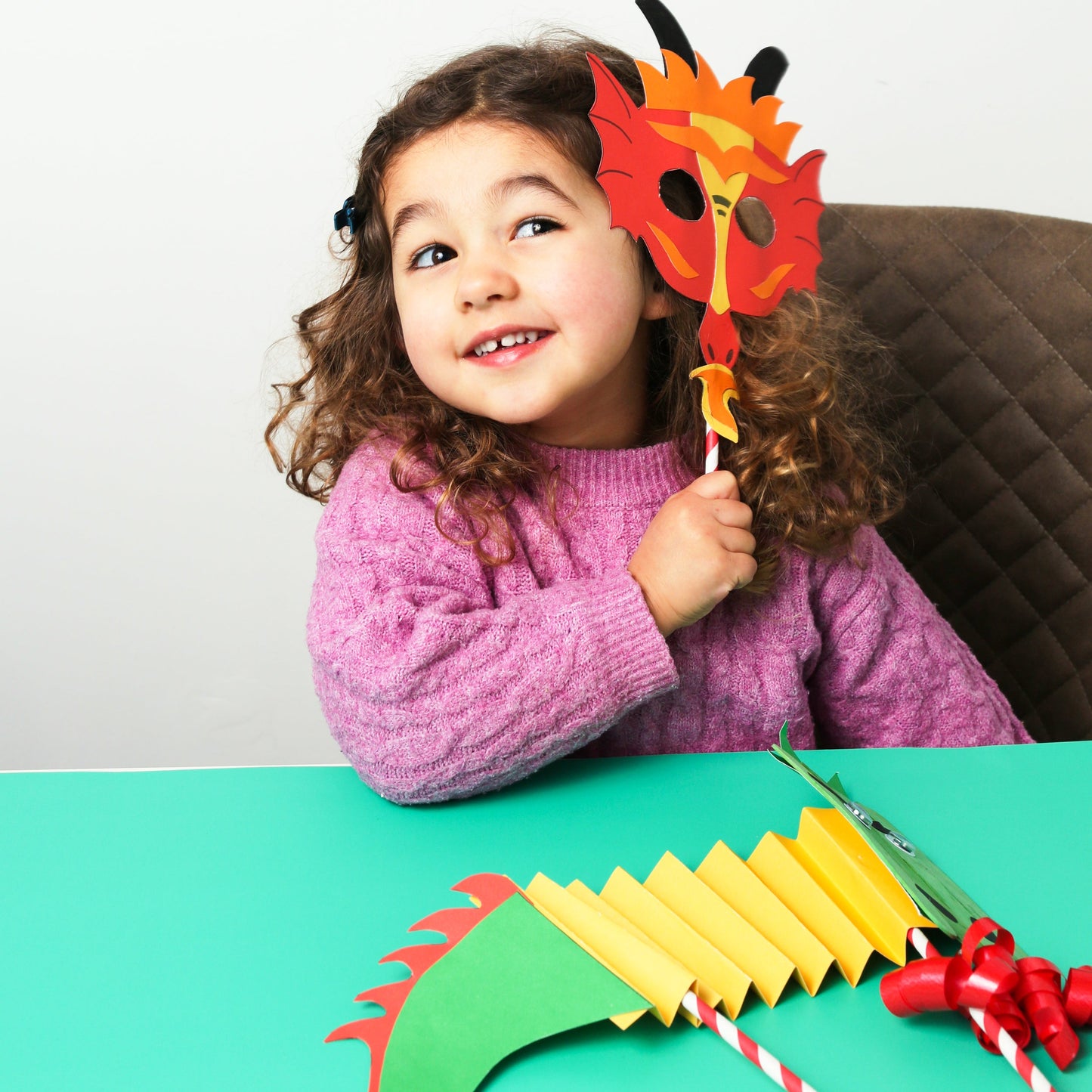 Child wearing a colorful dragon mask sitting at a table with craft materials.