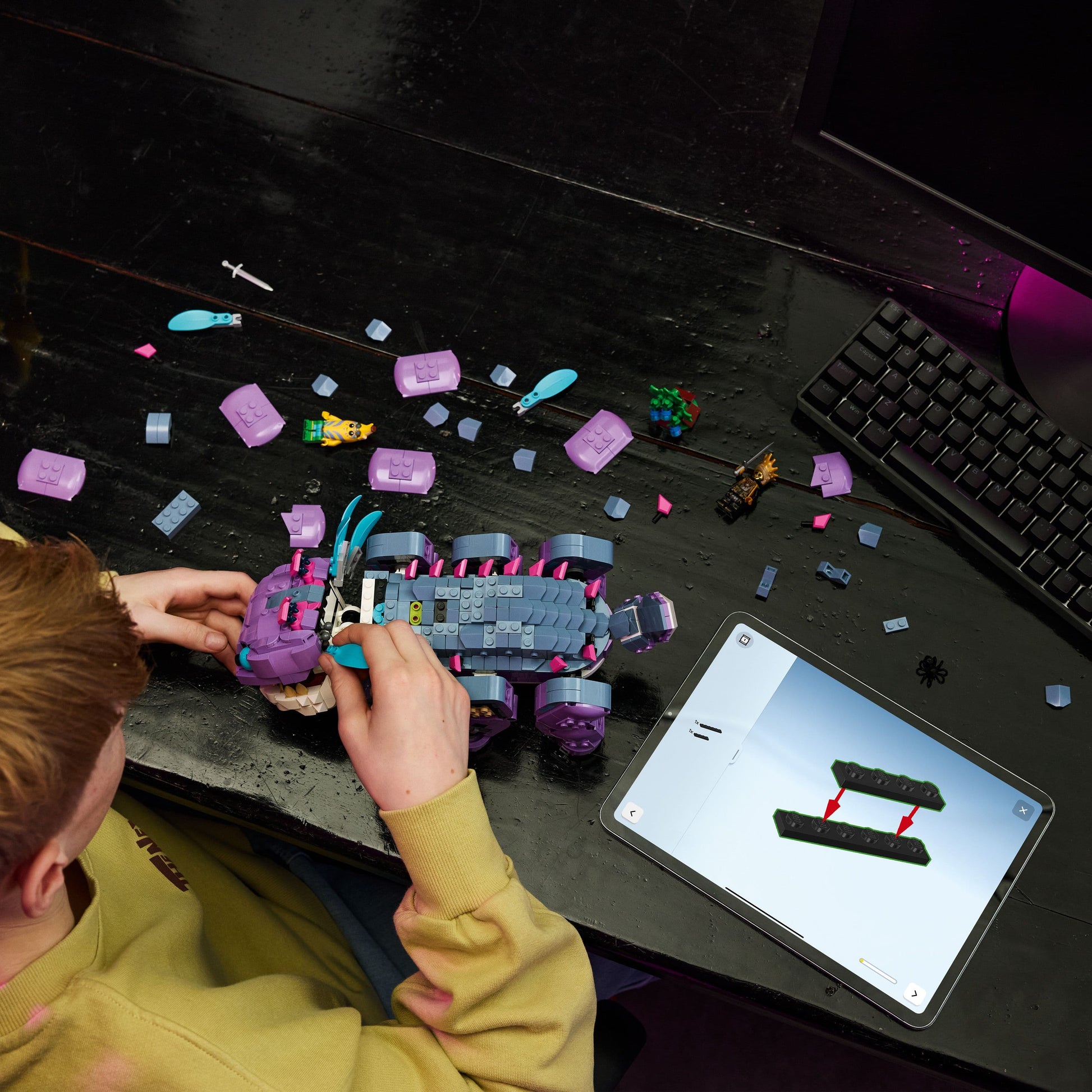 Person building a toy vehicle on a desk with a tablet displaying design images.